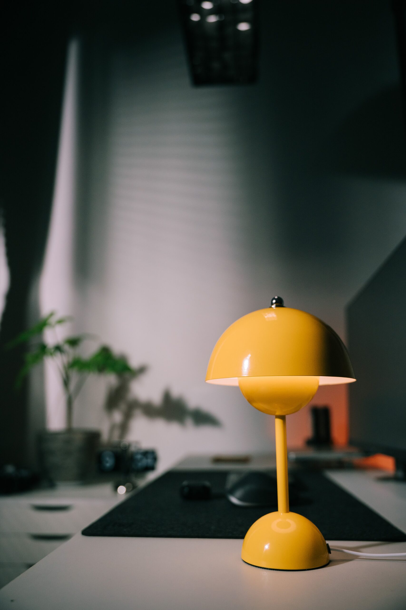 A bright yellow desk lamp illuminates a modern, minimalist workspace with a plant and shadows.